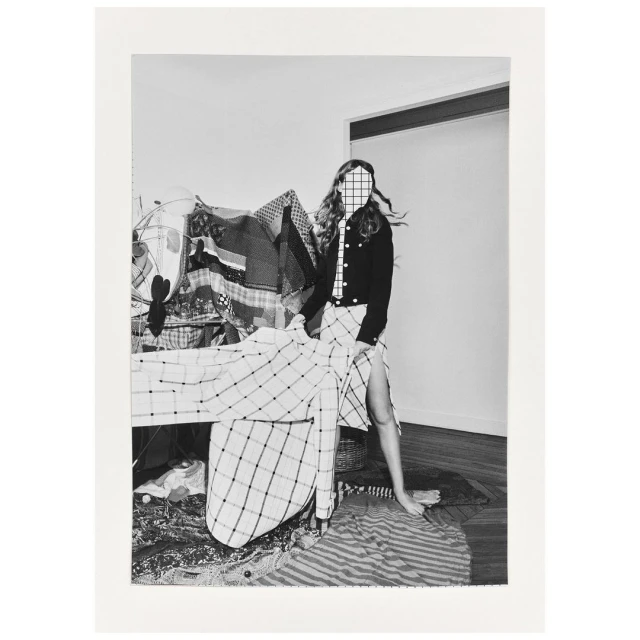 Monochrome portrait of a woman wearing a plaid skirt, jacket, and tie, posed indoors with clothing racks and patterned fabrics. 