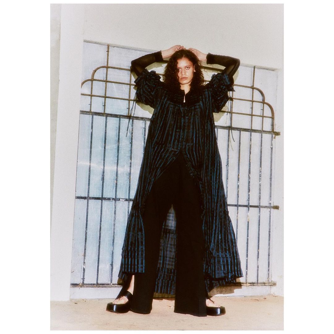 Model in dark patterned dress with hands behind head posing in front of a metal security gate, photographed by Adrian Samson