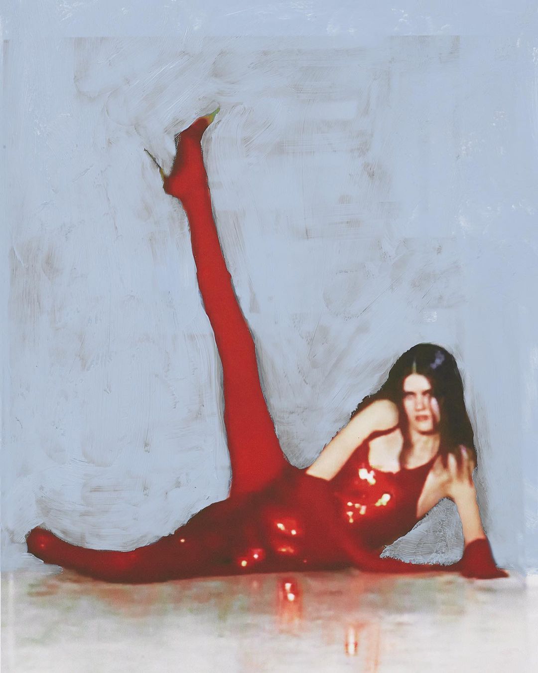 Woman lying down in a red, sequined bodysuit with matching heels and stockings, one leg raised, against a gray and white painted backdrop.