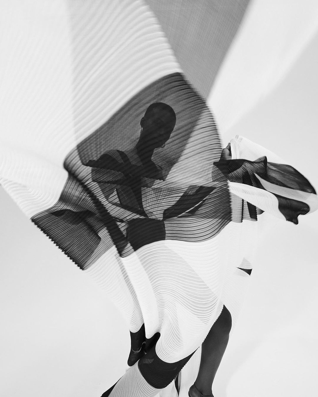 Monochrome fashion shot features an abstract view of a person draped in flowing, pleated fabric against a bright white backdrop in studio lighting.