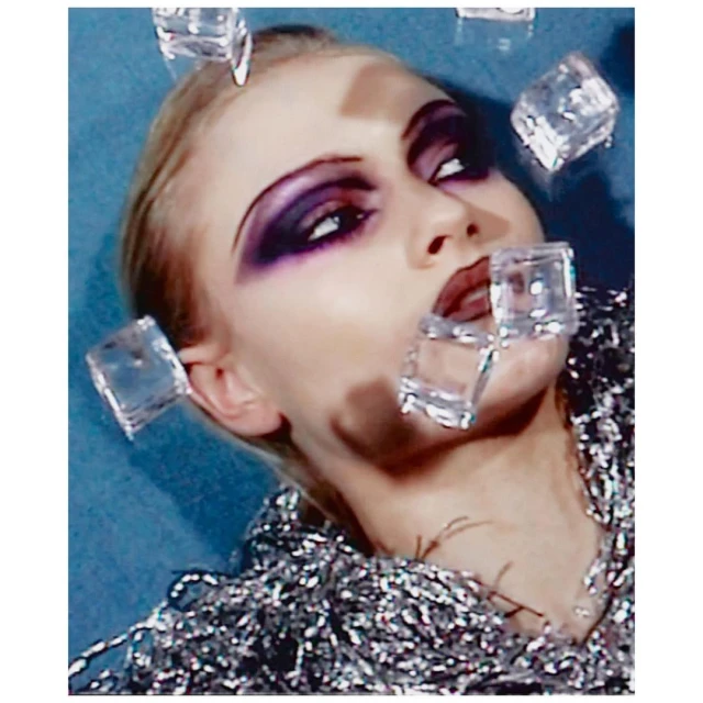 Close-up of a woman with dramatic makeup and shimmery collar, posing with several transparent ice cubes around her face and mouth.