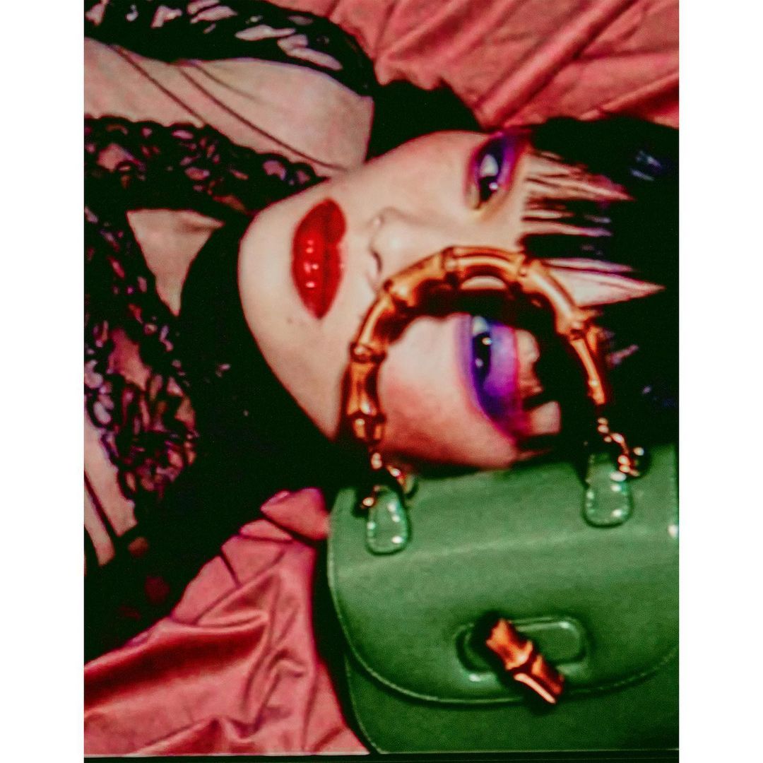 Female fashion model with bold makeup lies on pink satin, wearing a lace top and posing with a green Gucci bamboo-handle purse.