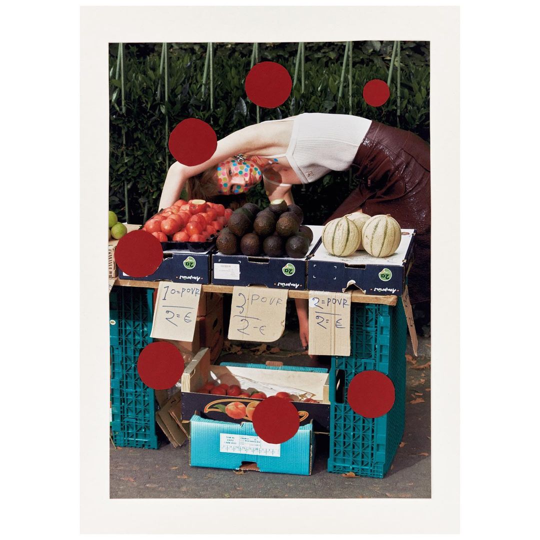 A person with colorful face paint is bending over a market stand filled with fruit against an outdoor, leafy green background.