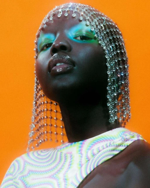Close-up of a woman with dark skin and blue eyeshadow, adorned with a jeweled headpiece, against an orange background in a portrait.