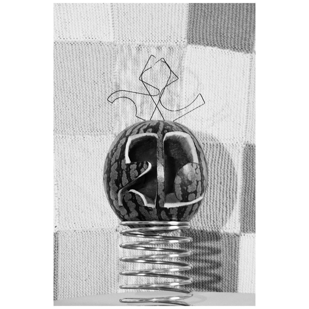 Black and white still life of a carved watermelon balanced on a metal spring against a square-patterned fabric backdrop.
