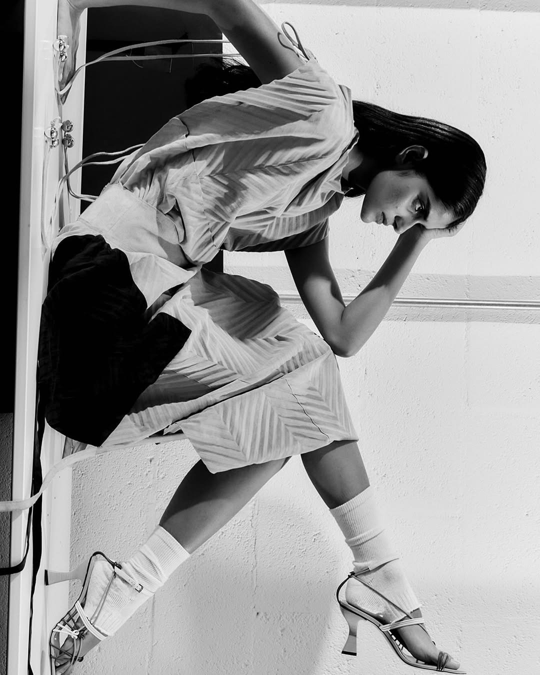 A young woman in a patterned dress and strappy heels poses inside a white doorway in a black and white fashion shot.