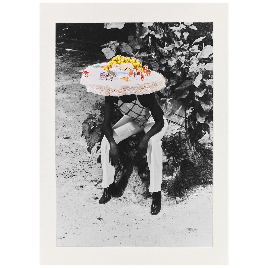 A person sits with a tablecloth on their head adorned with toy animals and yellow spheres, captured in a monochromatic outdoor setting with lush foliage.