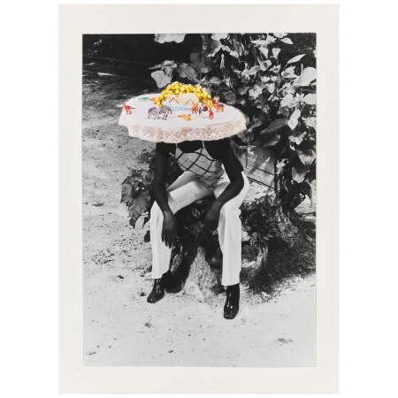 A person sits with a tablecloth on their head adorned with toy animals and yellow spheres, captured in a monochromatic outdoor setting with lush foliage.