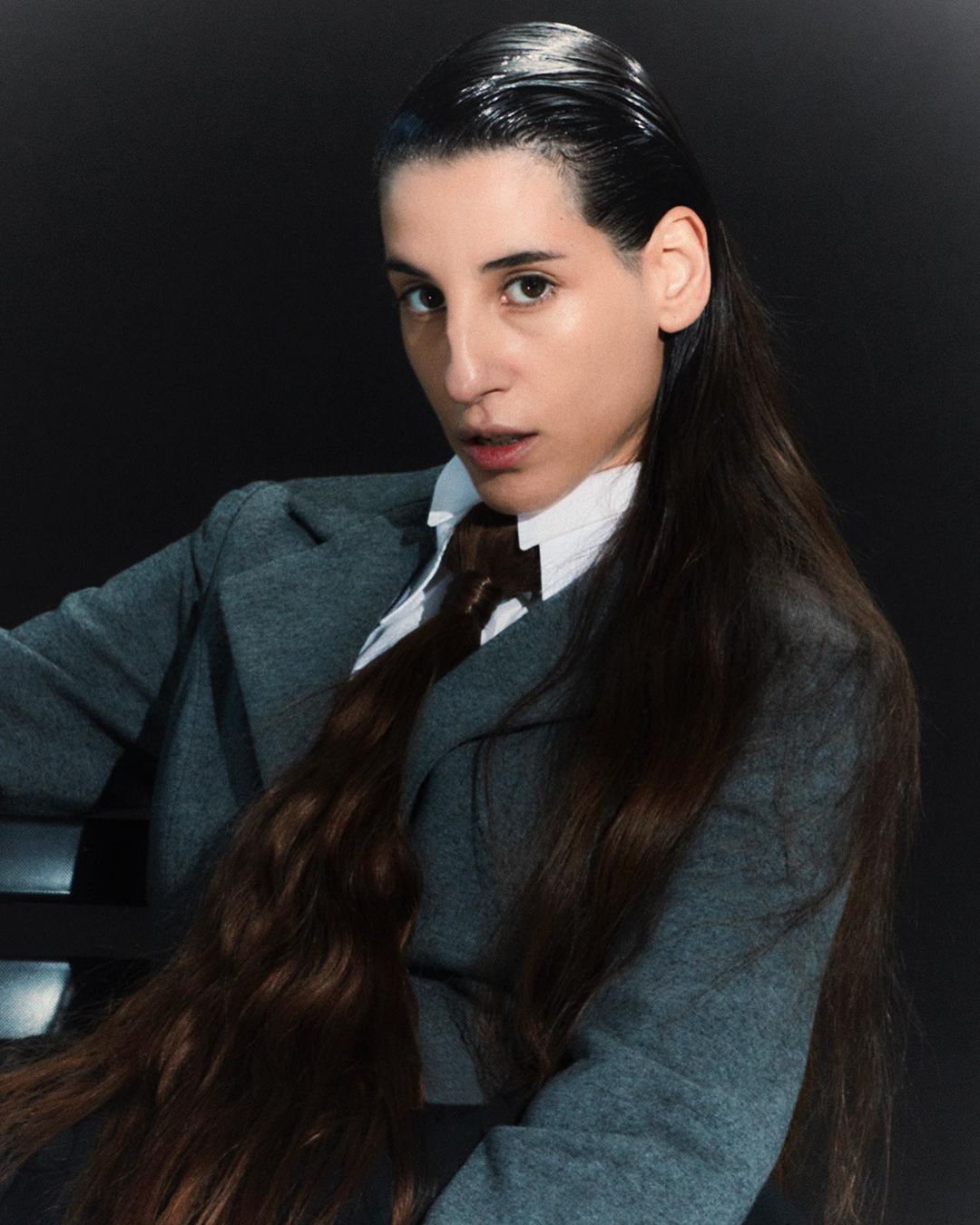 Portrait of a sleek-haired person in a grey suit with a tie against a dark backdrop, exuding a sophisticated androgynous vibe.