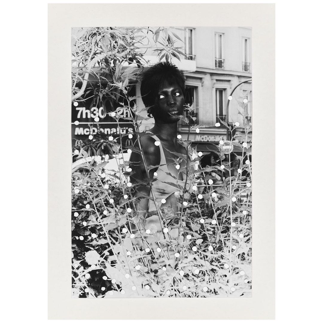 Black and white portrait of an African-American woman amidst foliage, with a glimpse of a Parisian street in the background.
