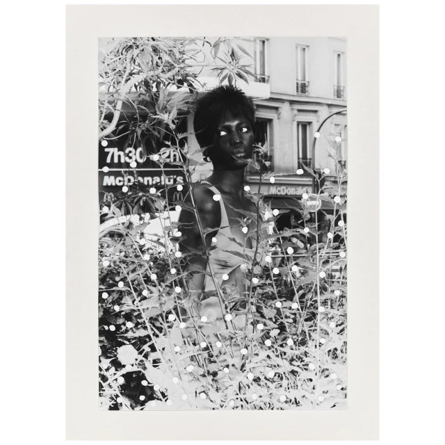 Black and white portrait of an African-American woman amidst foliage, with a glimpse of a Parisian street in the background.