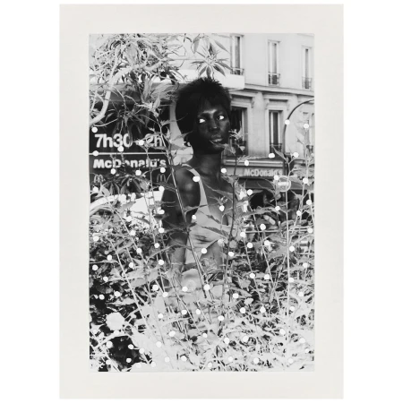 Black and white portrait of an African-American woman amidst foliage, with a glimpse of a Parisian street in the background.