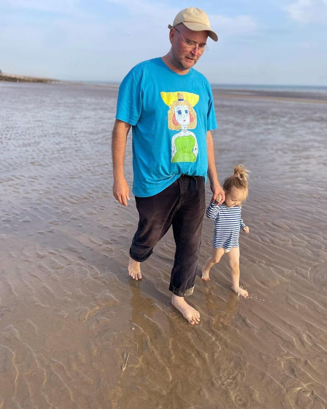 Man and toddler stroll barefoot on a sandy beach under a bright sky. The man wears a t-shirt with cartoon art.