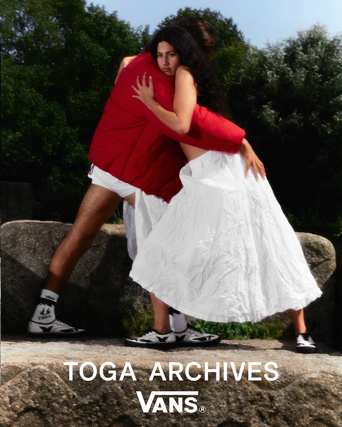 An adult couple embrace in an outdoor fashion shot featuring a man in a red jacket and a woman in a white dress near rocks and trees.