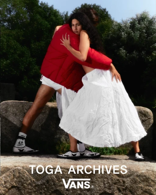 An adult couple embrace in an outdoor fashion shot featuring a man in a red jacket and a woman in a white dress near rocks and trees.