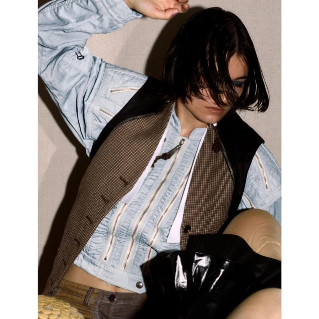 Stylish woman in a blue zippered jacket and plaid vest posing indoors with a serious facial expression, captured in medium shot.