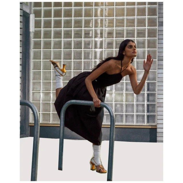 Woman in a black dress and yellow heels poses with a bicycle rack in an urban setting. She's holding onto the rack and extending her arm.