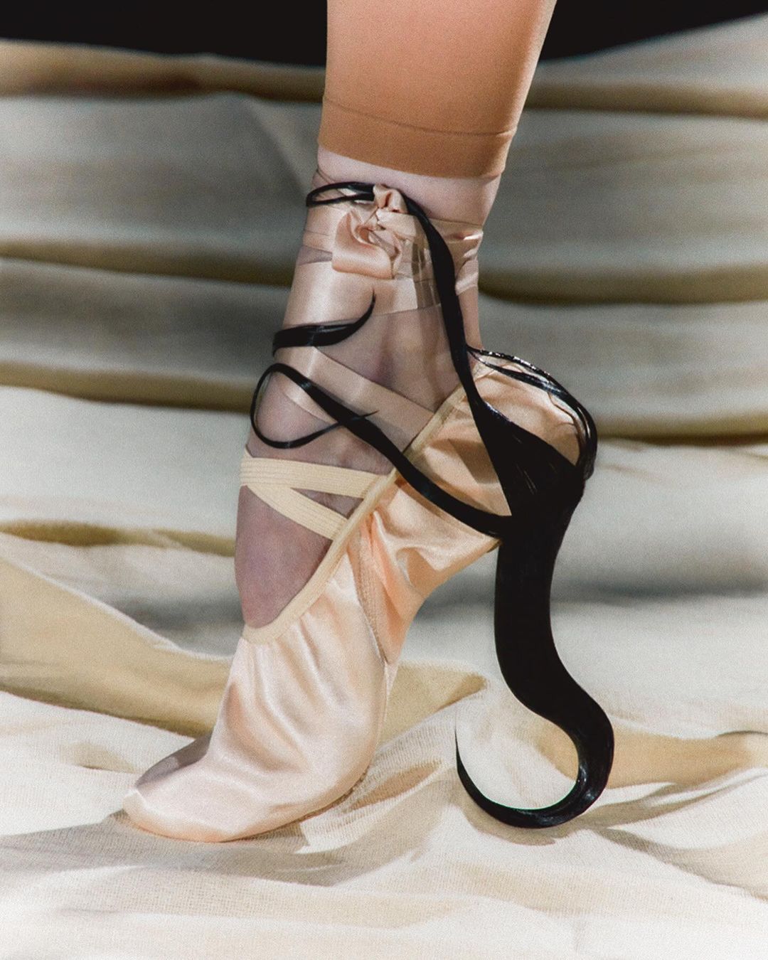 Close-up studio shot of a ballerina's foot wearing a pink pointe shoe with black ribbon detailing, against a cream fabric background.