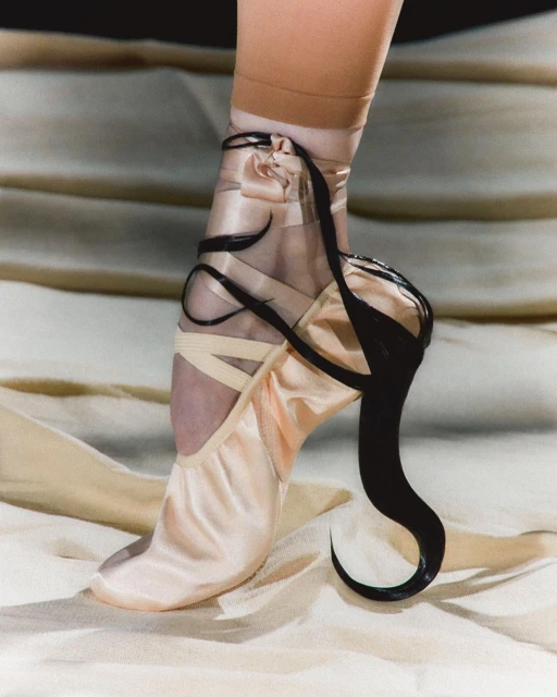 Close-up studio shot of a ballerina's foot wearing a pink pointe shoe with black ribbon detailing, against a cream fabric background.