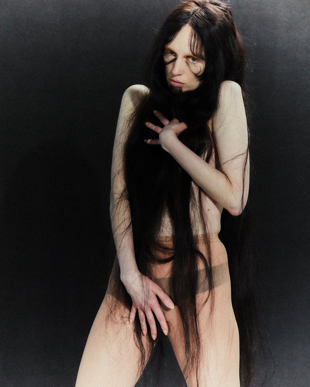 Adult female poses against a dark background with long dark hair, covering parts of her body, wearing neutral tights in a studio setting.