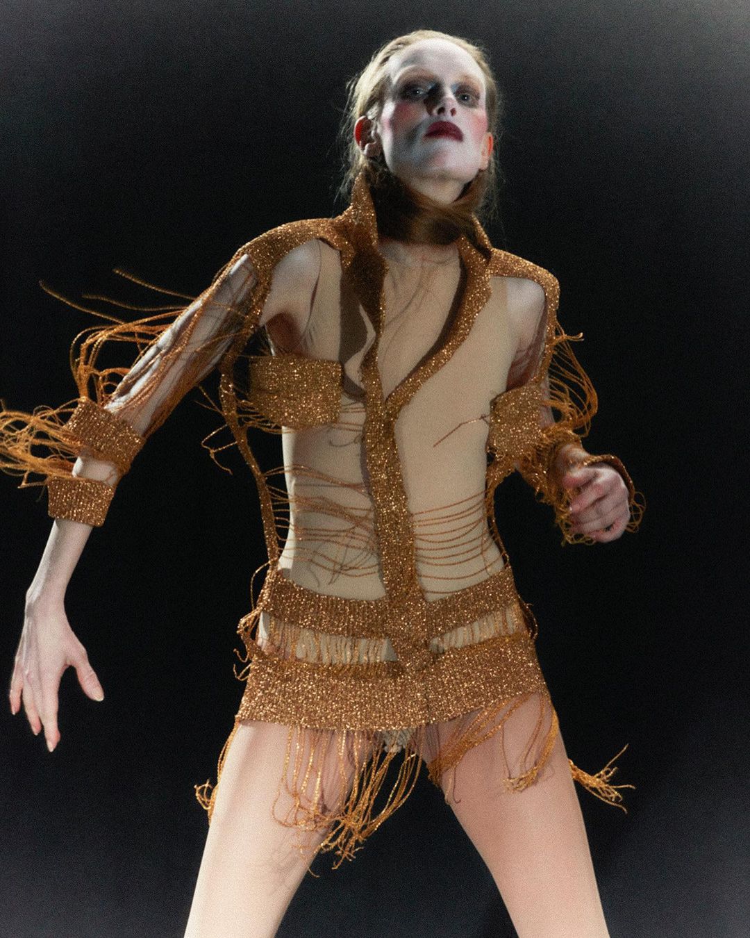 Fashion model with pale skin and dark lipstick walks a dark runway in a copper-colored fringed shirt, shorts, and tan-colored bodysuit.