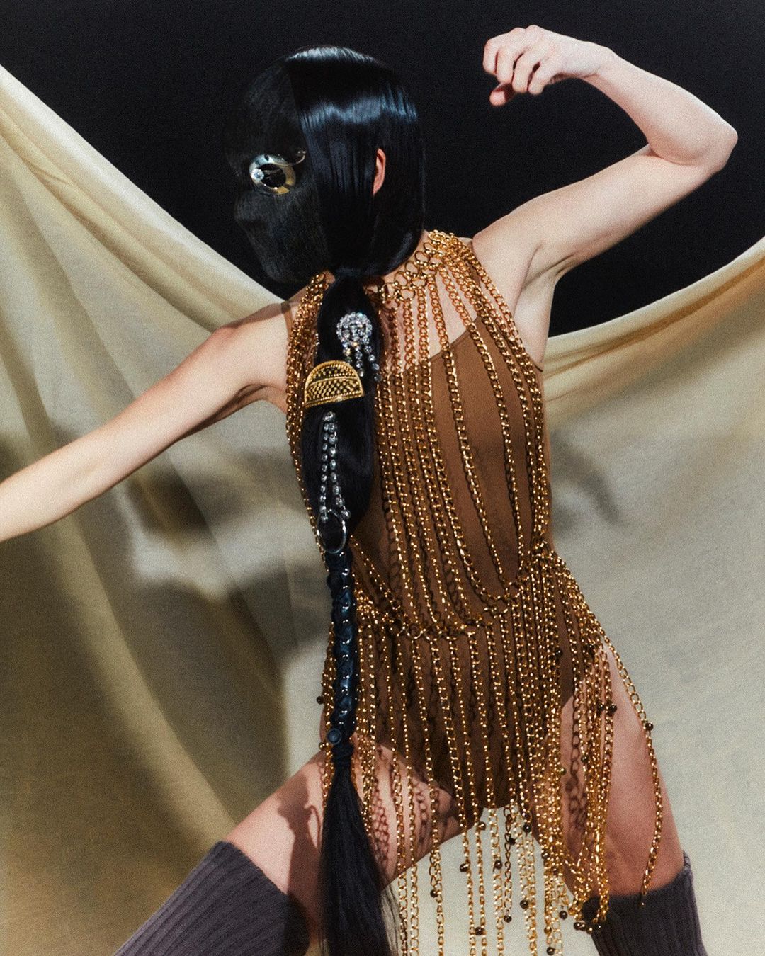 A single woman in a full black face covering poses in a tan dress of draped gold chains against a simple black studio backdrop.