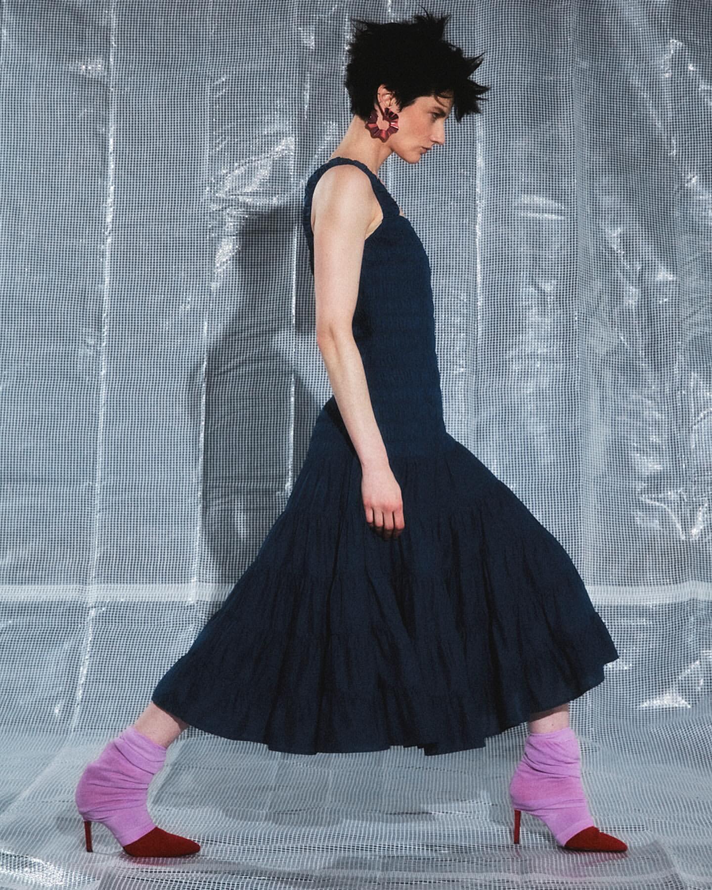Woman in a navy dress and lavender and burgundy heels poses in front of a silver patterned backdrop in a fashion forward photo.