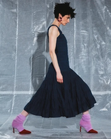 Woman in a navy dress and lavender and burgundy heels poses in front of a silver patterned backdrop in a fashion forward photo.