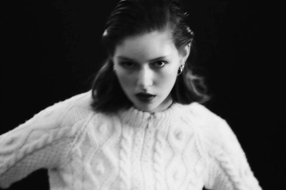 Black and white portrait of a young adult woman wearing a white cable-knit sweater against a dark background, looking directly at the camera.