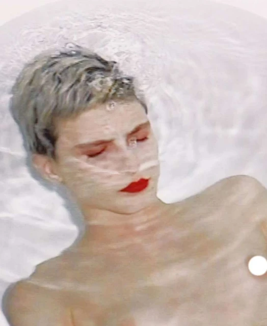 Adult female with closed eyes submerged in water, red lipstick, short blonde hair, pale skin, inside bright white bathtub.