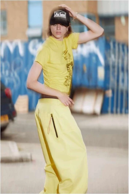 A young adult female poses in a yellow tee and pants in an urban setting, wearing a black baseball cap with the Aries logo.