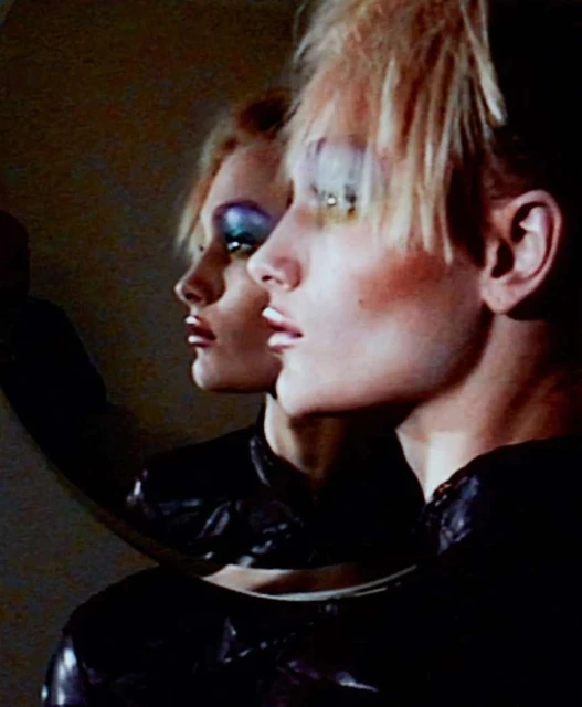Portrait of a person in profile with dramatic makeup reflected in a mirror, glossy black top, and neutral background with soft lighting.