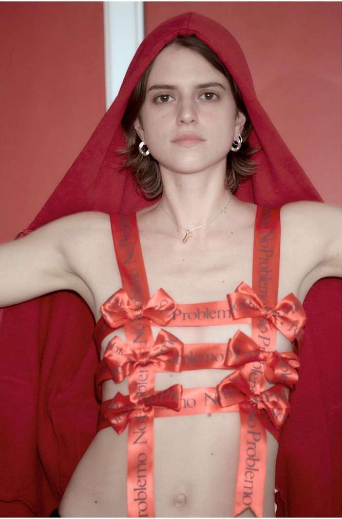 Woman in red hood and decorative ribbon top poses against a red background in a studio shot with soft lighting and neutral expression.