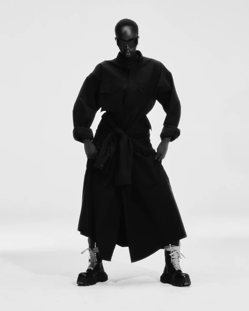 Woman in dark clothing and chunky boots stands in a brightly lit photo studio, posing for a fashion shot in black and white.
