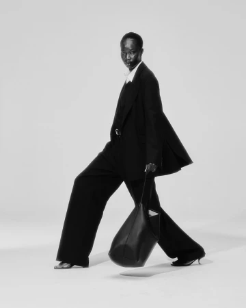Black and white editorial photograph of a model in a dark suit walking and holding a large leather tote bag.