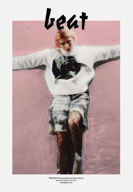 Editorial fashion shot of a pink-haired model wearing a printed sweater and sequined shorts against a pink backdrop.