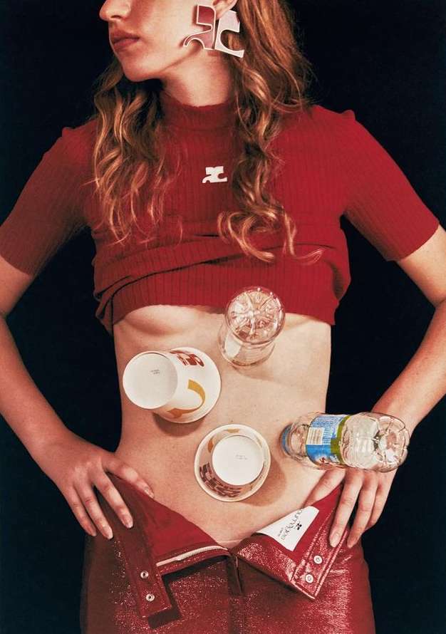 Fashion editorial shot of a model in a red top and pants with glasses and cups balanced on her abdomen, set against a dark background.