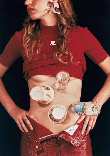 Fashion editorial shot of a model in a red top and pants with glasses and cups balanced on her abdomen, set against a dark background.