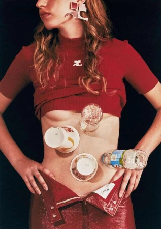 Fashion editorial shot of a model in a red top and pants with glasses and cups balanced on her abdomen, set against a dark background.