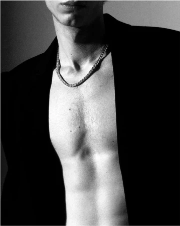 Close-up monochrome portrait of a man wearing an open dark jacket and a chain necklace against a light background.