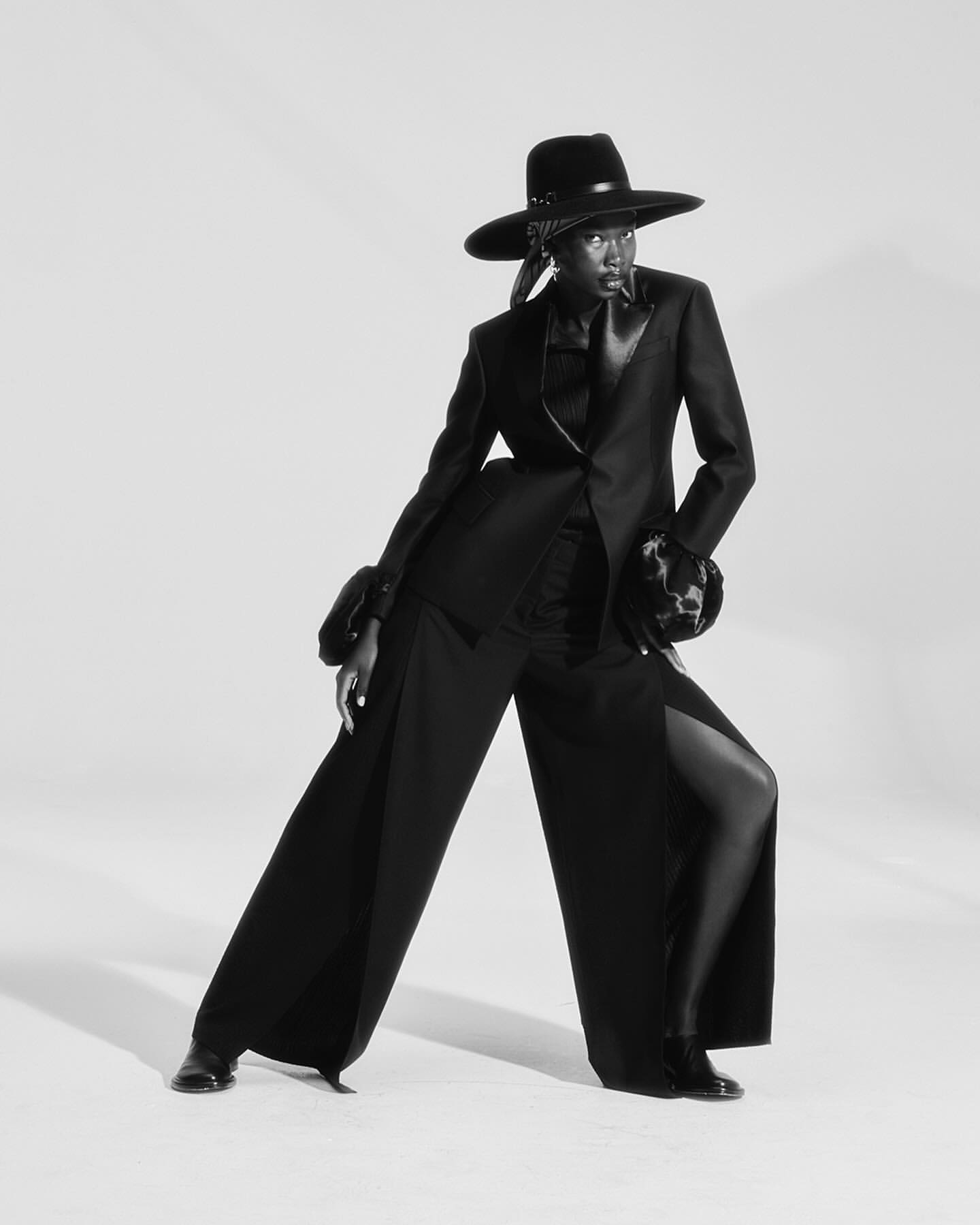 Monochrome fashion portrait of a slender adult female in a black suit and wide-brimmed hat, posing against a plain white background.