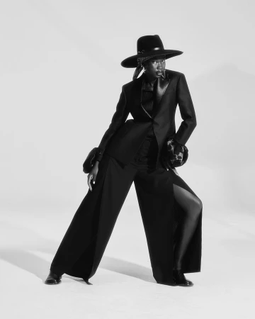 Monochrome fashion portrait of a slender adult female in a black suit and wide-brimmed hat, posing against a plain white background.