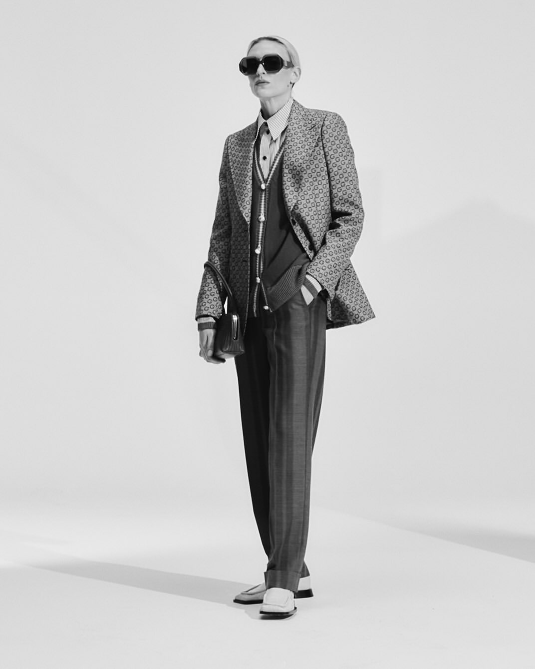 Model in patterned blazer and dark trousers holding a handbag against a seamless white background.