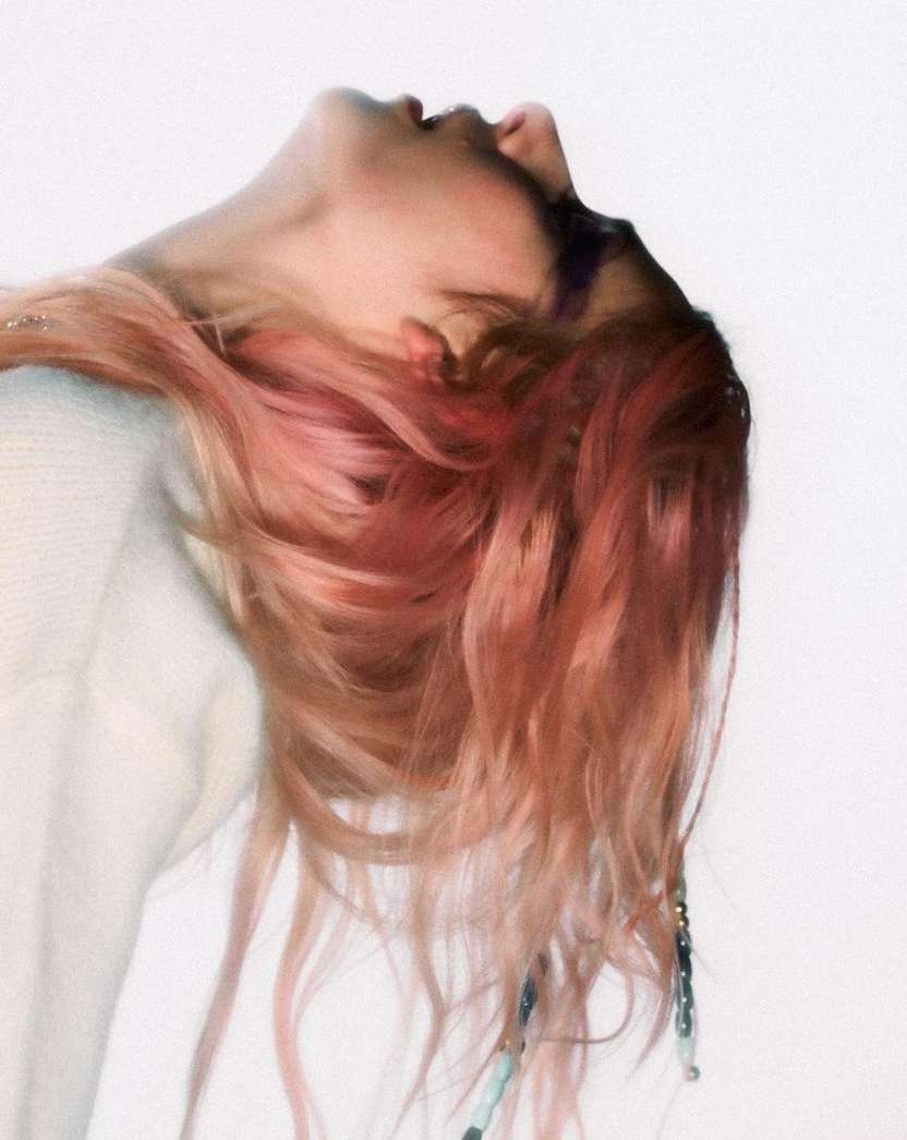 Headshot of a young adult female with light skin and light pink hair tilted back, wearing an off-white sweater and accessories in her hair.