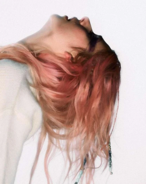 Headshot of a young adult female with light skin and light pink hair tilted back, wearing an off-white sweater and accessories in her hair.