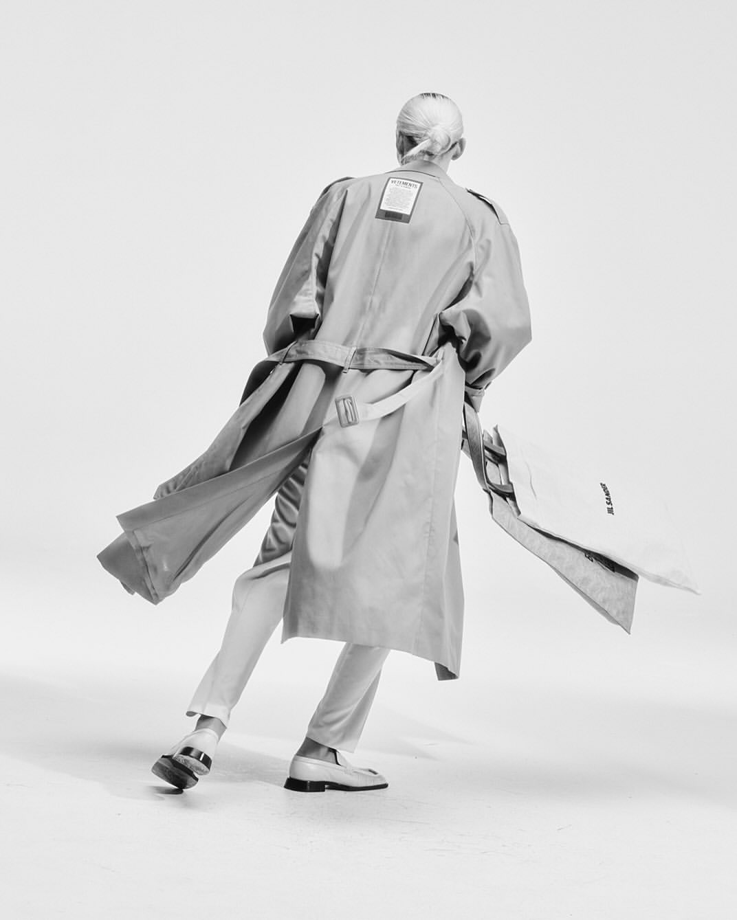 Back view of a woman in a trench coat, pants, and loafers, holding a garment bag against a white backdrop in a studio setting.