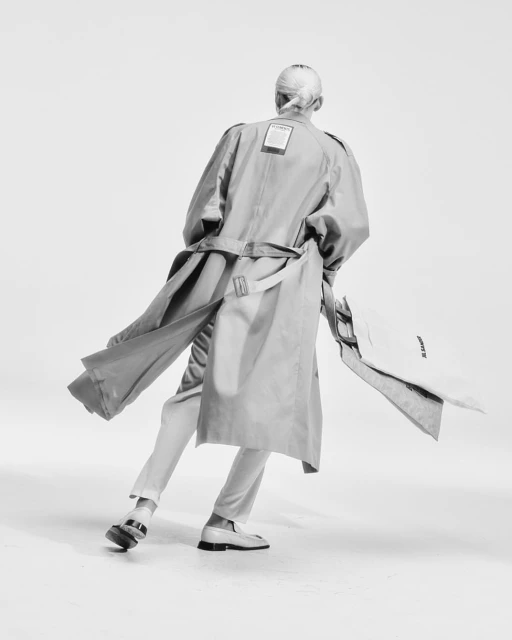 Back view of a woman in a trench coat, pants, and loafers, holding a garment bag against a white backdrop in a studio setting.
