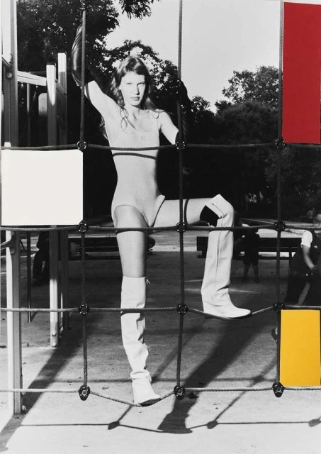 Fashion shot of a slender adult woman posing in a tan bodysuit and white thigh-high boots climbing on playground equipment in a park setting.