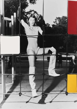 Fashion shot of a slender adult woman posing in a tan bodysuit and white thigh-high boots climbing on playground equipment in a park setting.
