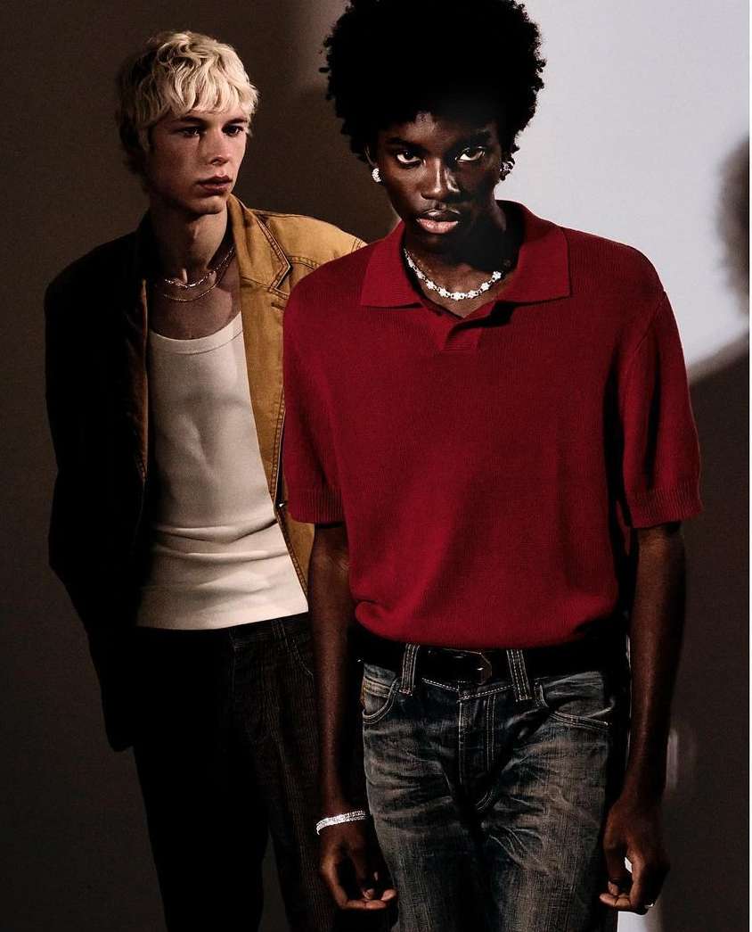 Two stylish men in casual attire pose indoors; one in a red polo shirt and jeans, the other in a white tank top and tan jacket.